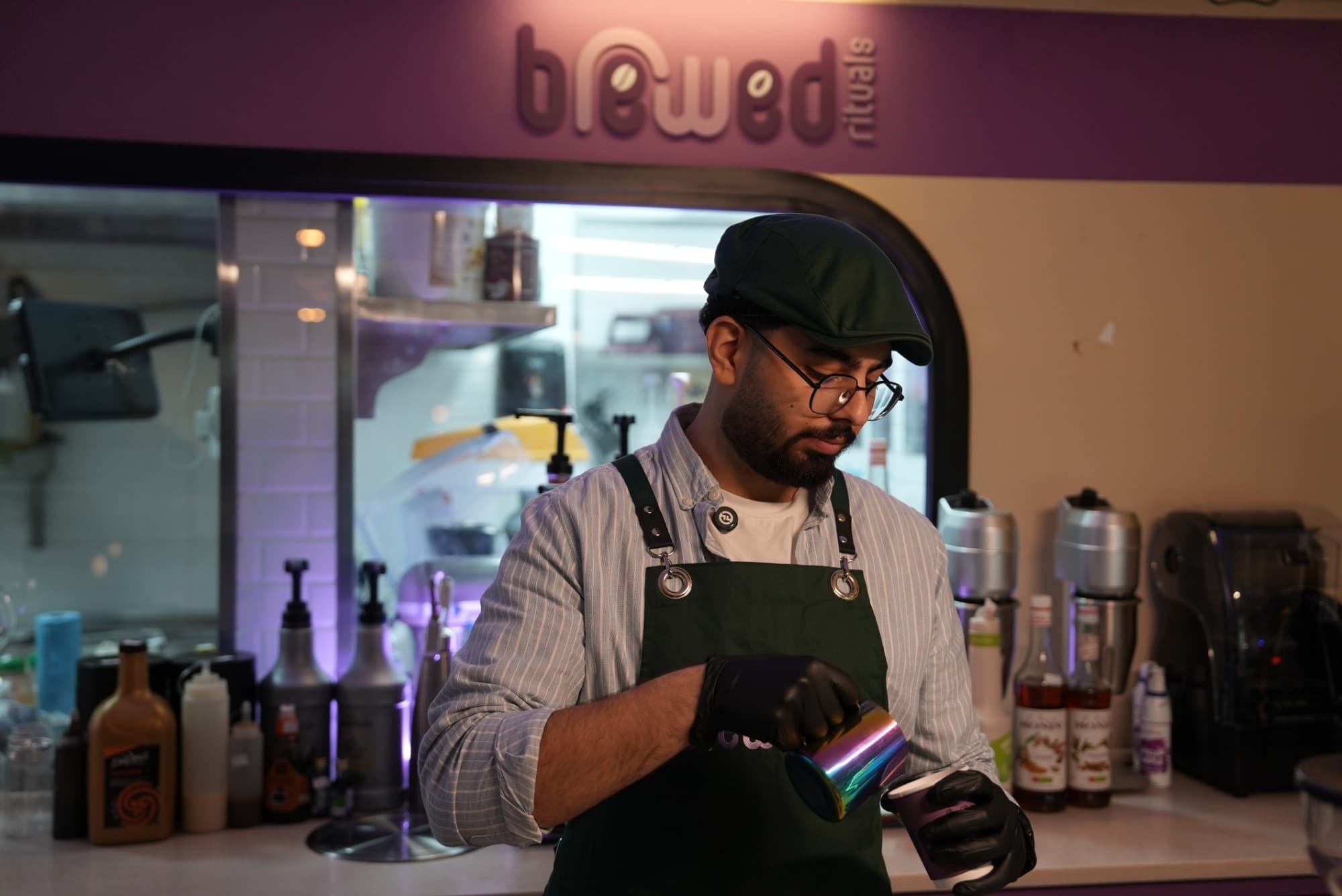 A Brewed Rituals barista pouring steamed milk beneath the shop's sign