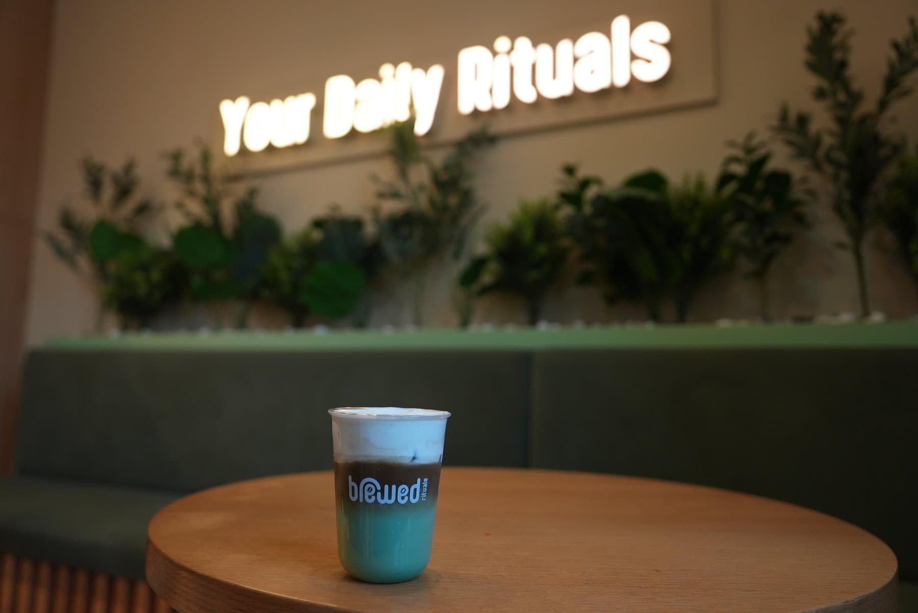 A drink in a Brewed cup on a café table under the 'Your Daily Rituals' sign
