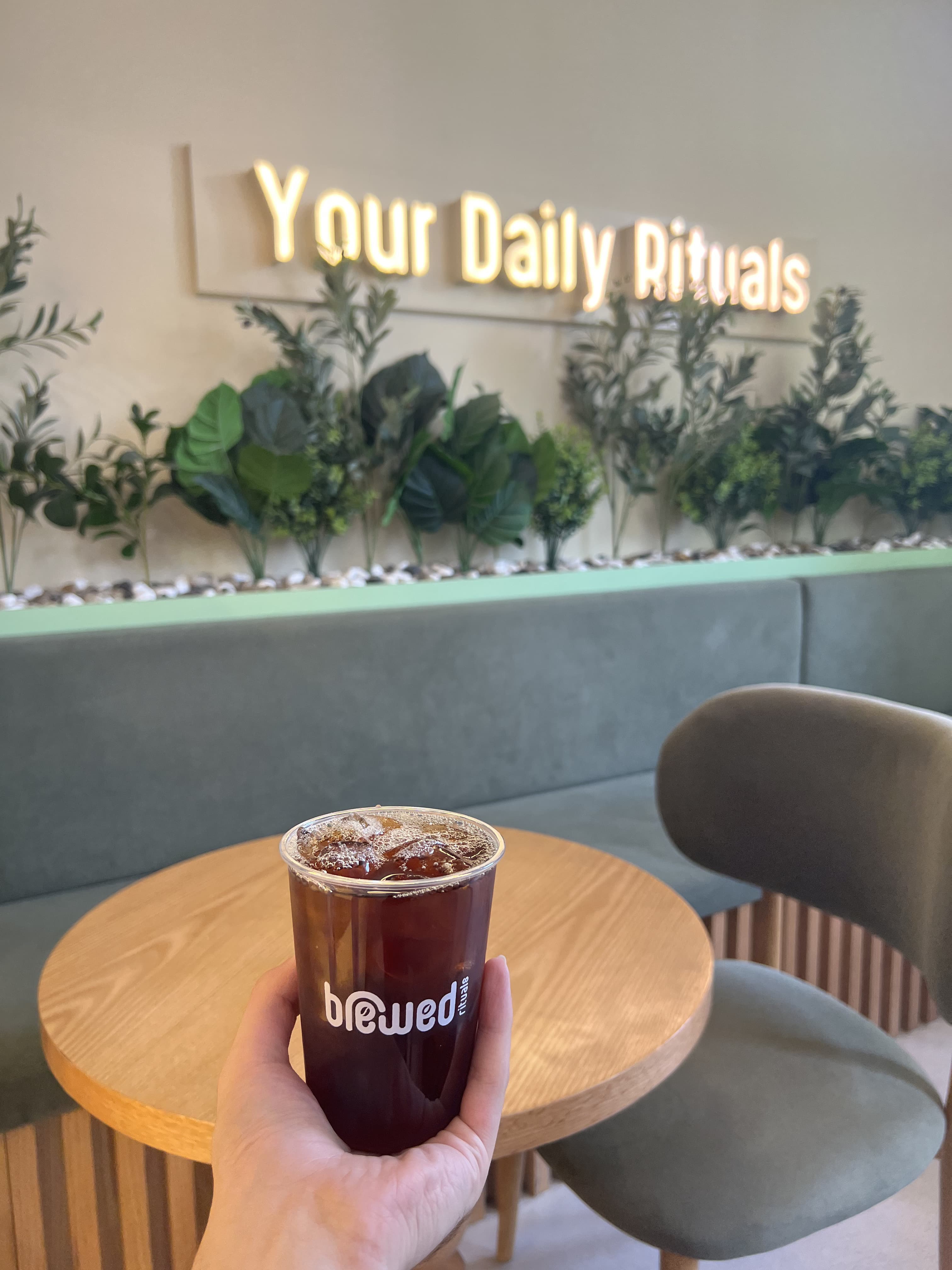A guest holding an iced Brewed Rituals coffee in front of the 'Your Daily Rituals' sign